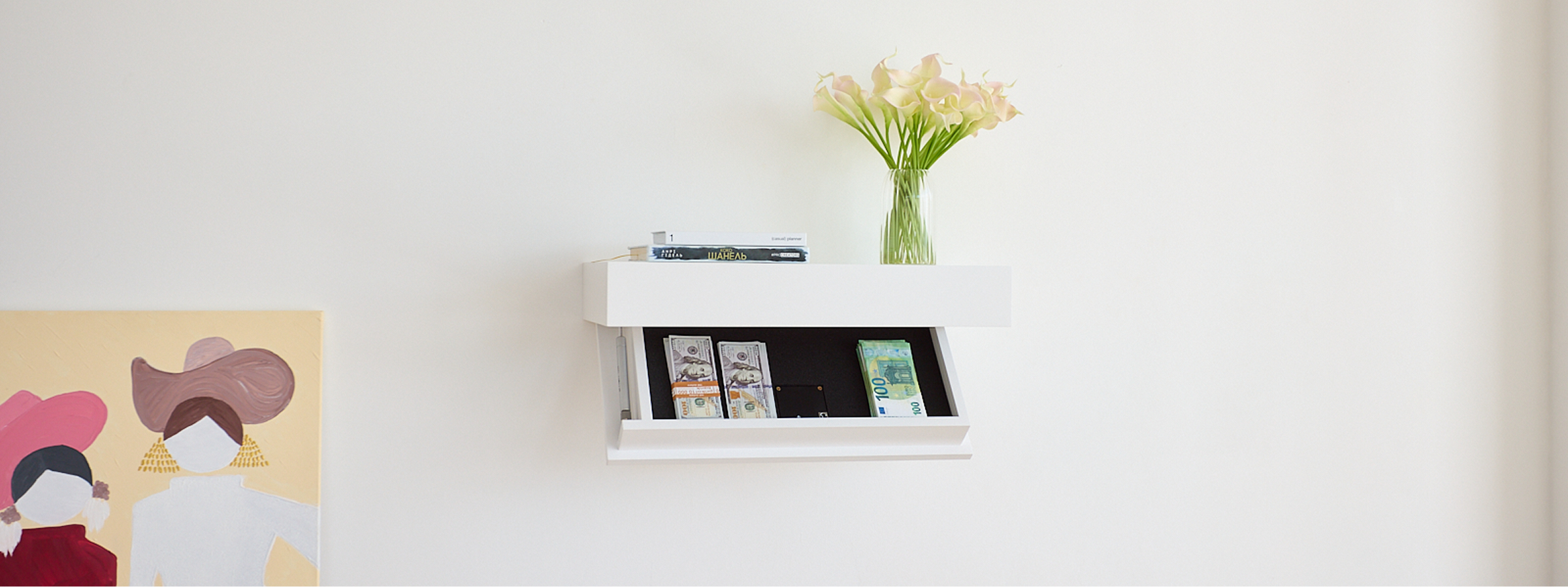 A white wall-mounted floating shelf with an open secret lower drawer, styled with a vase of pale yellow flowers and a few books or cards, against a plain white wall