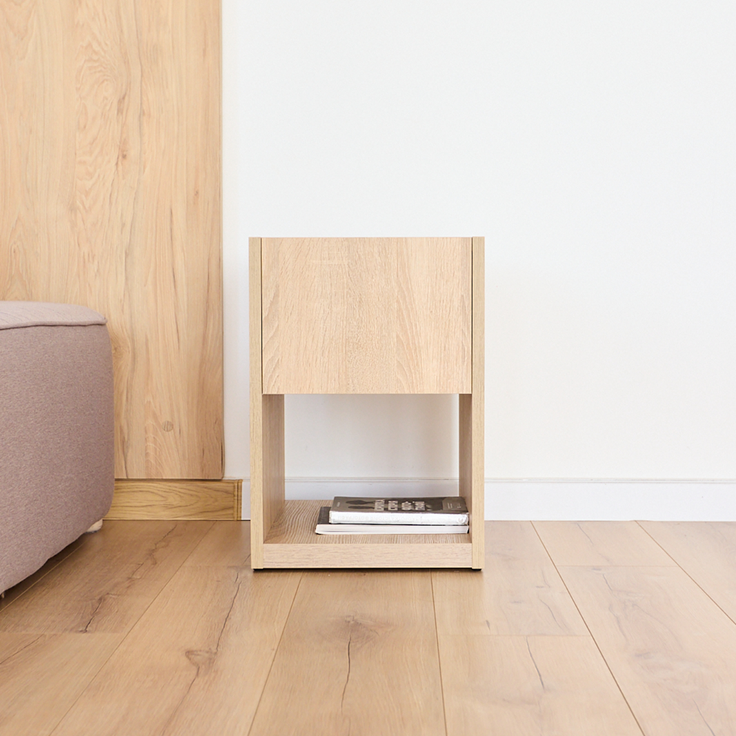 Minimalist nightstand with top-access hidden RFID-locked compartment by Hiddo. A light natural oak side table with an open lower shelf, styled in a minimalist room setting with a white wall, wooden floor, and a pink ottoman partially visible on the left.