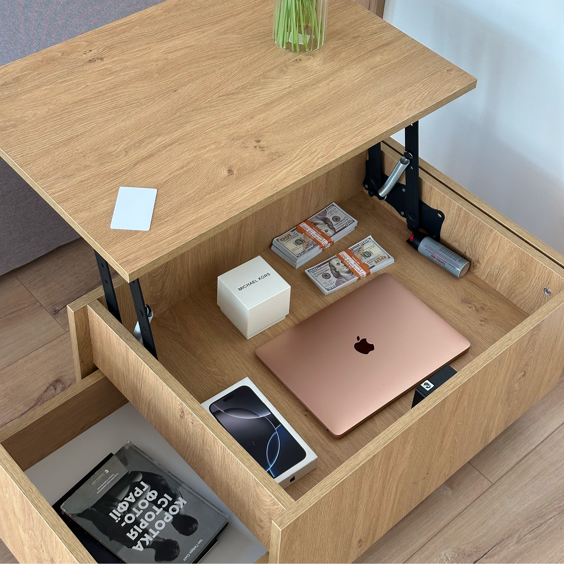 A light oak wood lift-top storage table with an open drawer beneath the desktop, shown holding a rose gold laptop, phone, cash, and other items. The table is photographed in a real room with wood flooring and a vase on top.