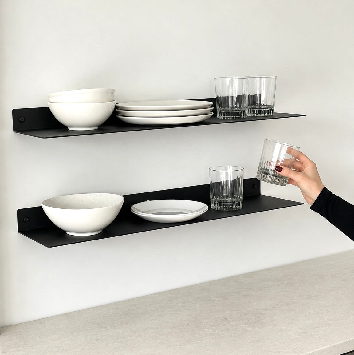 Two black floating wall shelves mounted on a white wall above a light stone countertop, styled with white bowls, stacked plates, and clear drinking glasses. A person's hand is holding one of the clear glasses.