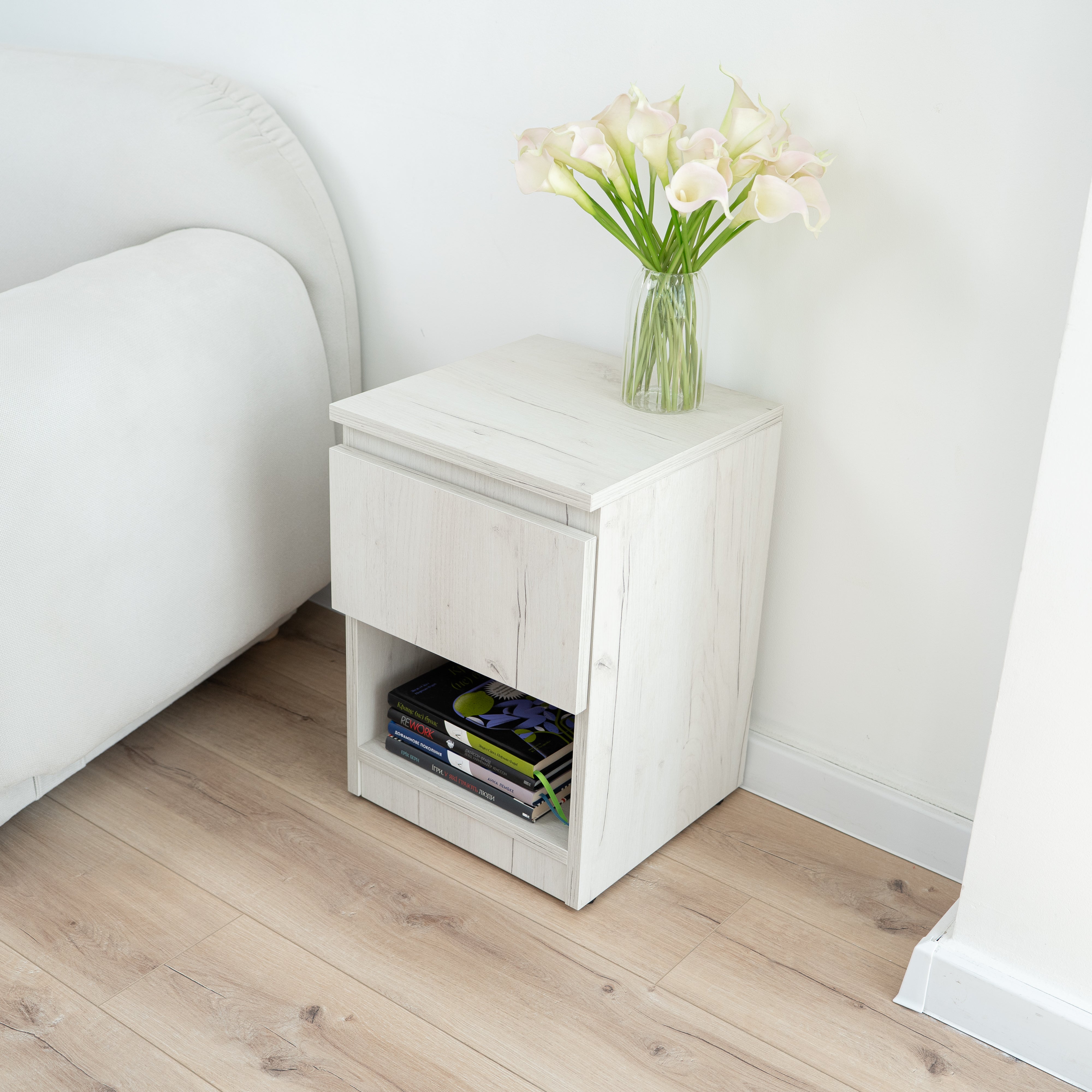 Narrow nightstand with two hidden compartments and RFID lock by Hiddo. A small white nightstand with a single drawer and open lower shelf, styled beside a bed in a bright bedroom. A clear glass vase with pale calla lilies sits on top, and books are stored in the open shelf.
