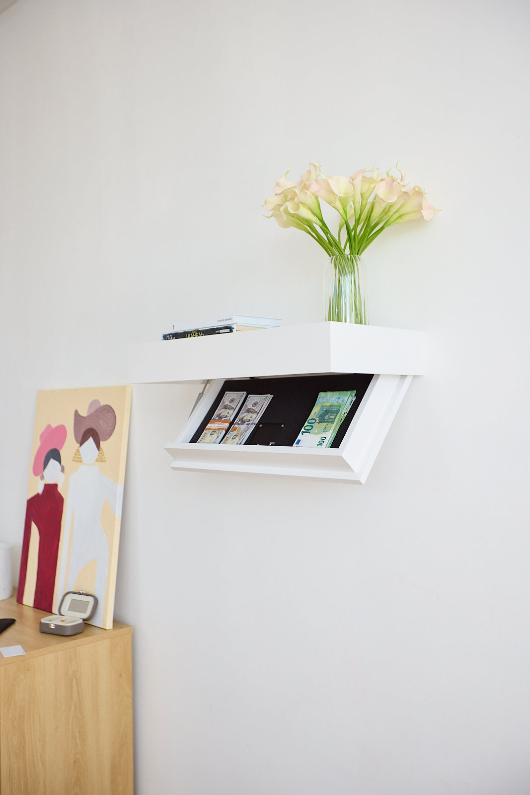 A white floating wall shelf with opened secret drawer mounted on a white wall, holding a clear glass vase with pale yellow flowers and a stack of books or magazines