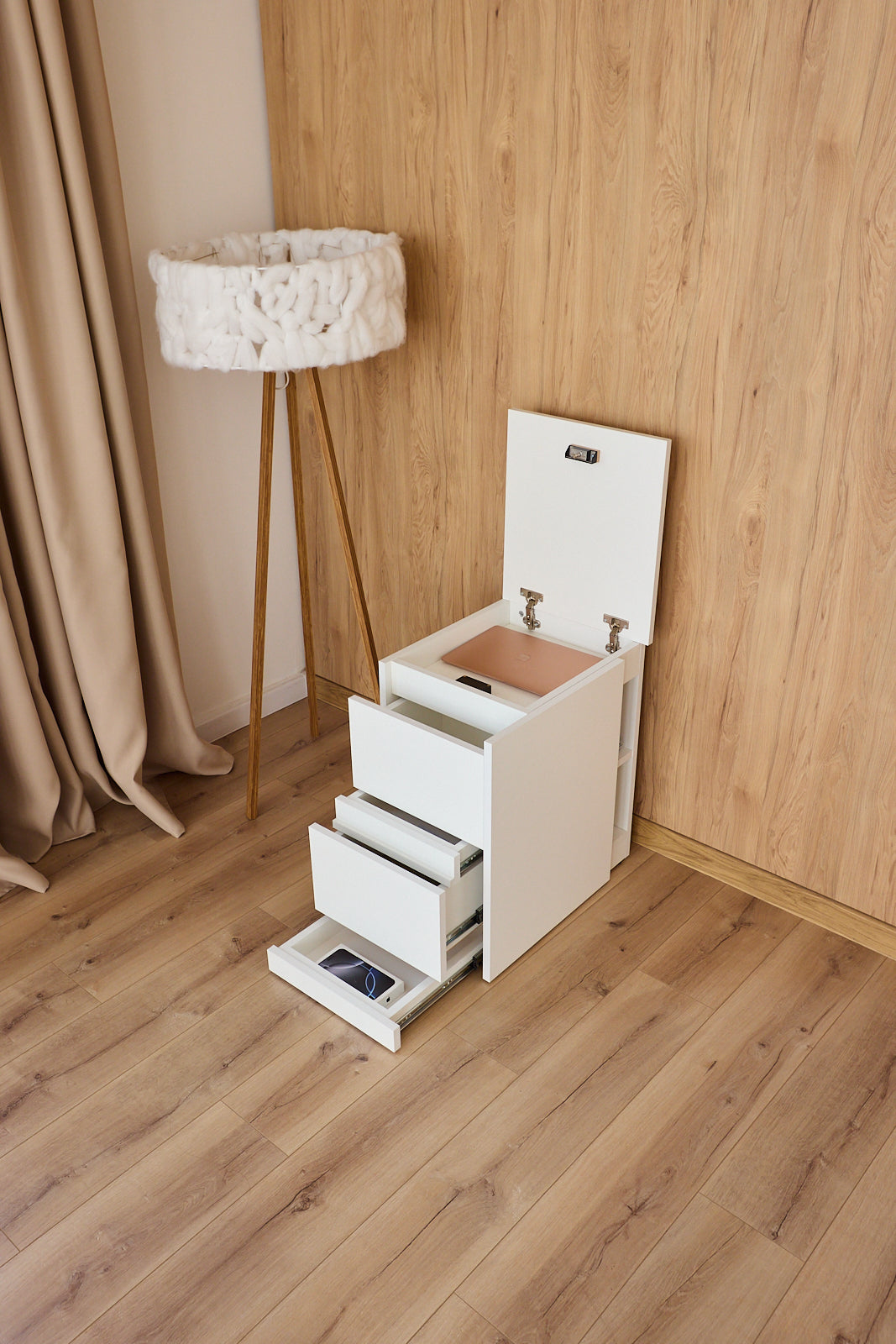 A white under-bed storage cabinet with three pull-out drawers and a lift-up lid, shown in a bedroom setting with wood flooring and a wooden wall paneling