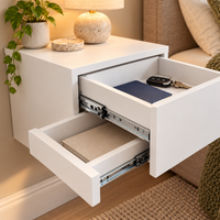 A modern white floating nightstand drawer shown in a real bedroom setting, with two drawers pulled open to reveal metal drawer slides. The top surface holds a potted plant, a lamp base, and a coaster, and the open drawers contain a blue notebook and keys.