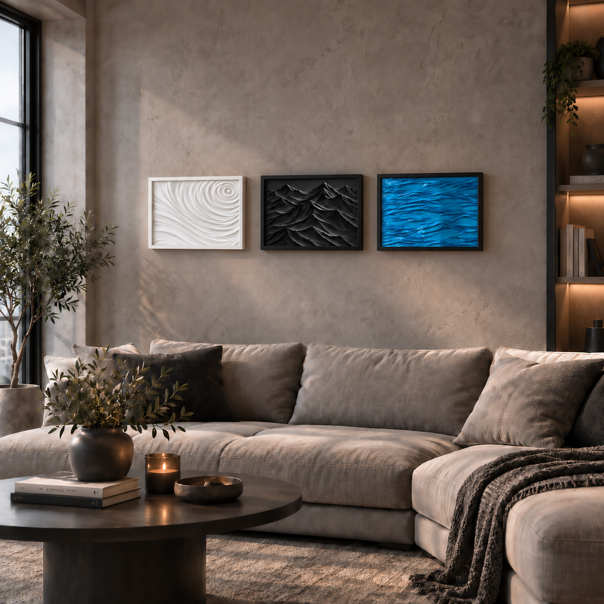 Three framed wall art prints hang above a beige sectional sofa in a modern living room. The prints show abstract wave-like textures in white, black, and vivid blue, with a textured concrete wall and a dark round coffee table in the foreground.