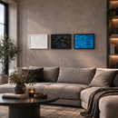 Three framed wall art prints hang above a beige sectional sofa in a modern living room. The prints show abstract wave-like textures in white, black, and vivid blue, with a textured concrete wall and a dark round coffee table in the foreground.