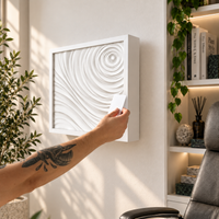 A white square wall-mounted art piece with a 3D swirling wave design is shown on a light wall, with a tattooed arm holding a small card in front of it. A bookshelf with plants and decor is visible on the right.