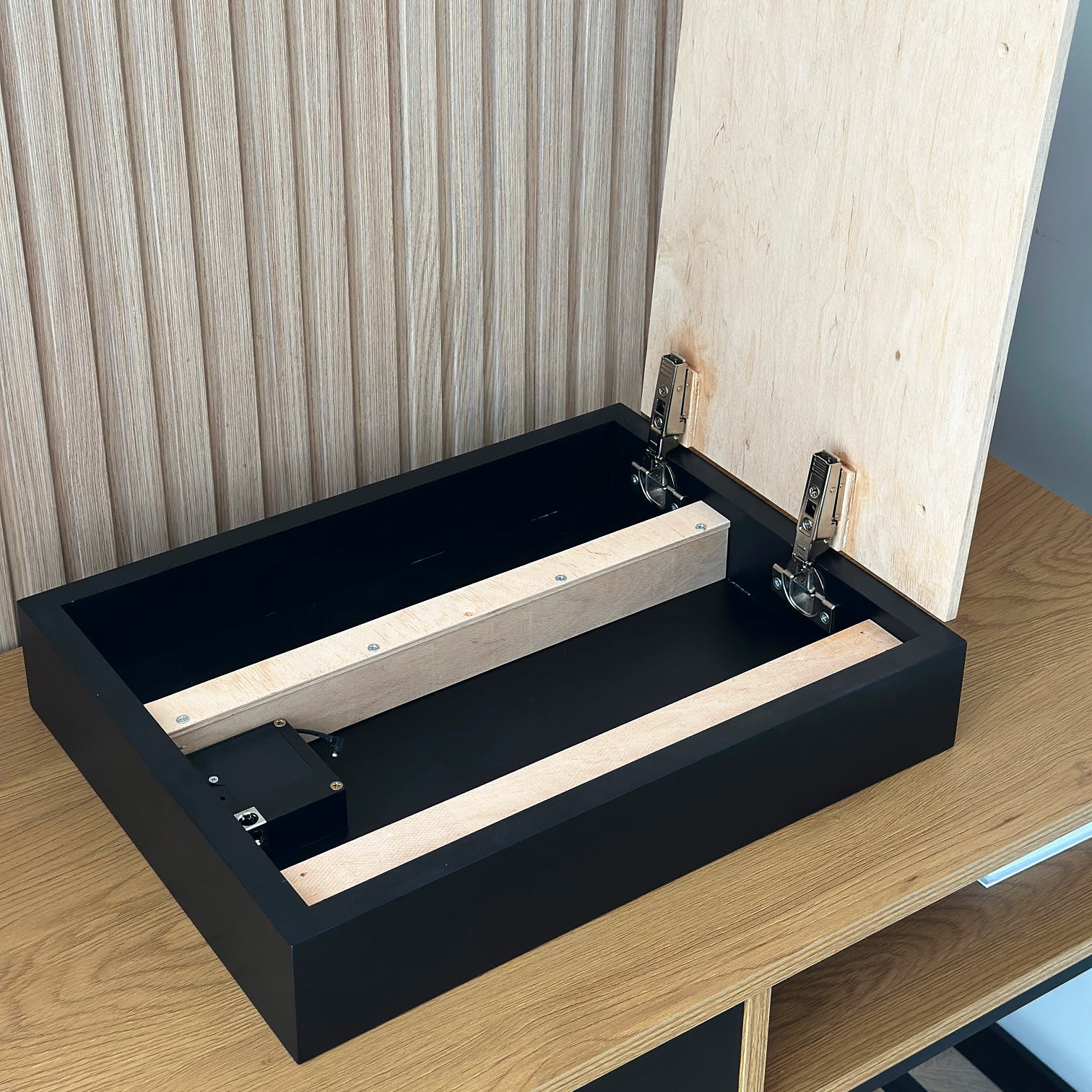 Hidden Wall Storage with Contactless RFID Lock. A black wooden storage box with its lid raised, showing the hinge hardware and light wood interior supports, placed on a wooden table against a paneled wall.