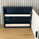 Modern Wave Wall Art with Concealed Storage. A black wall-mounted storage cabinet shown open, with the door swung to the right and light wood backing visible. The cabinet is photographed indoors against a slatted wood wall above a wood tabletop.