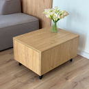 A rectangular wooden coffee table with a light oak finish and black legs, styled in a living room setting with a vase of white calla lilies on top.