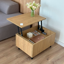 A light wood lift-top side table shown in a real room setting, with the top panel raised to reveal an open storage compartment underneath. The table is styled with a vase of white flowers on top and contains a laptop and other items inside the storage space.