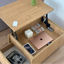 A light oak wood lift-top storage table with an open drawer beneath the desktop, shown holding a rose gold laptop, phone, cash, and other items. The table is photographed in a real room with wood flooring and a vase on top.