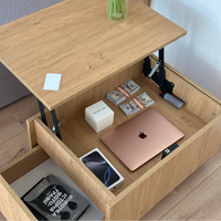 A light oak wood lift-top storage table with an open drawer beneath the desktop, shown holding a rose gold laptop, phone, cash, and other items. The table is photographed in a real room with wood flooring and a vase on top.
