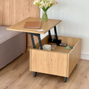 A light oak wood lift-top end table shown in a home setting, with its table top raised to show the black metal hydraulic lift mechanism. The table is styled with a brown notebook and a vase of white flowers, positioned next to a gray sofa on a light wood floor.