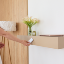 A beige floating wall shelf mounted on a white wall, styled with a glass vase of white calla lilies, a small candle, and a closed laptop. A person's arm and hand are visible on the left, holding a small white card near the shelf.