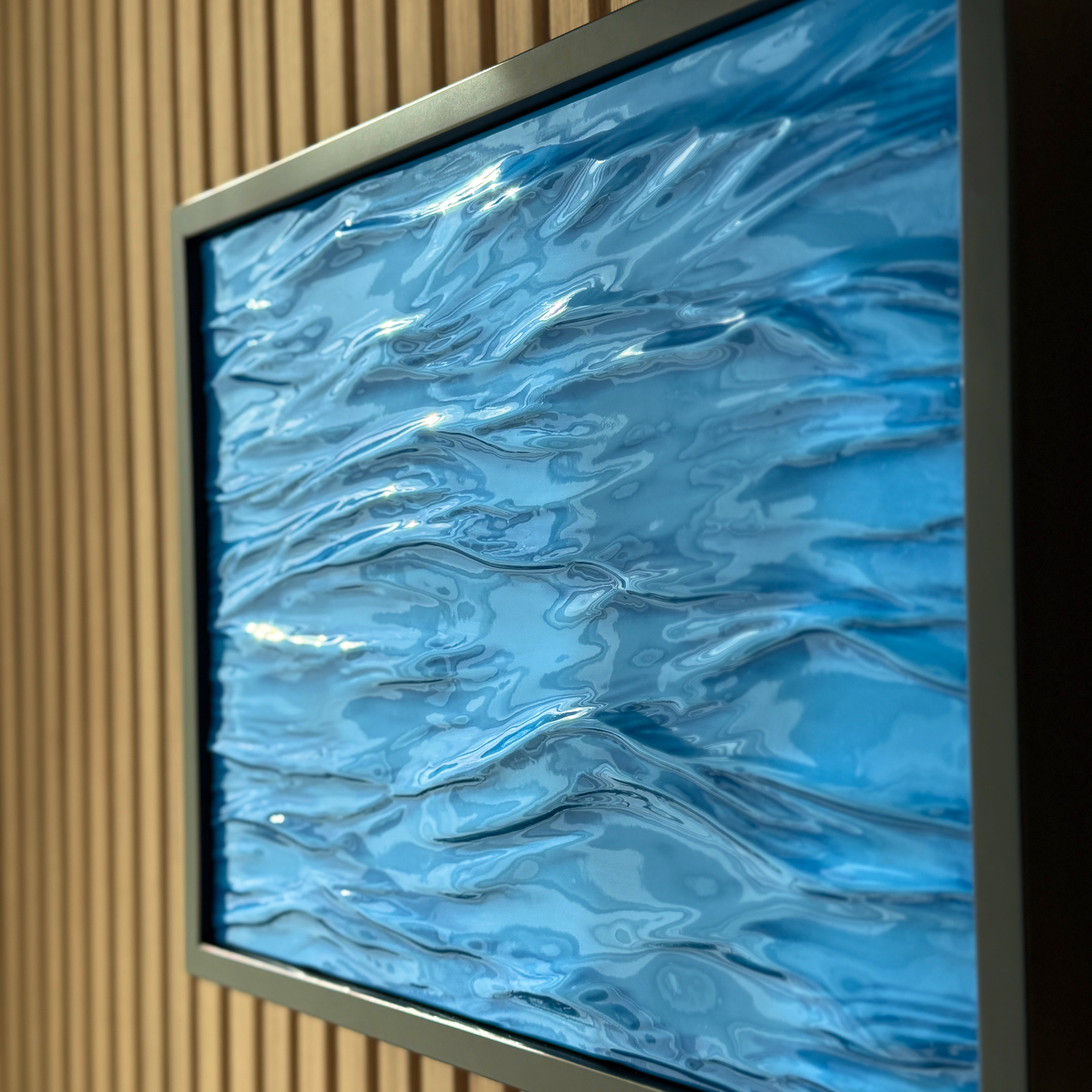 Modern Wave Wall Art with Concealed Storage. A square framed wall art piece featuring a glossy, textured blue abstract design resembling rippling water or liquid waves, mounted on a slatted wood wall.