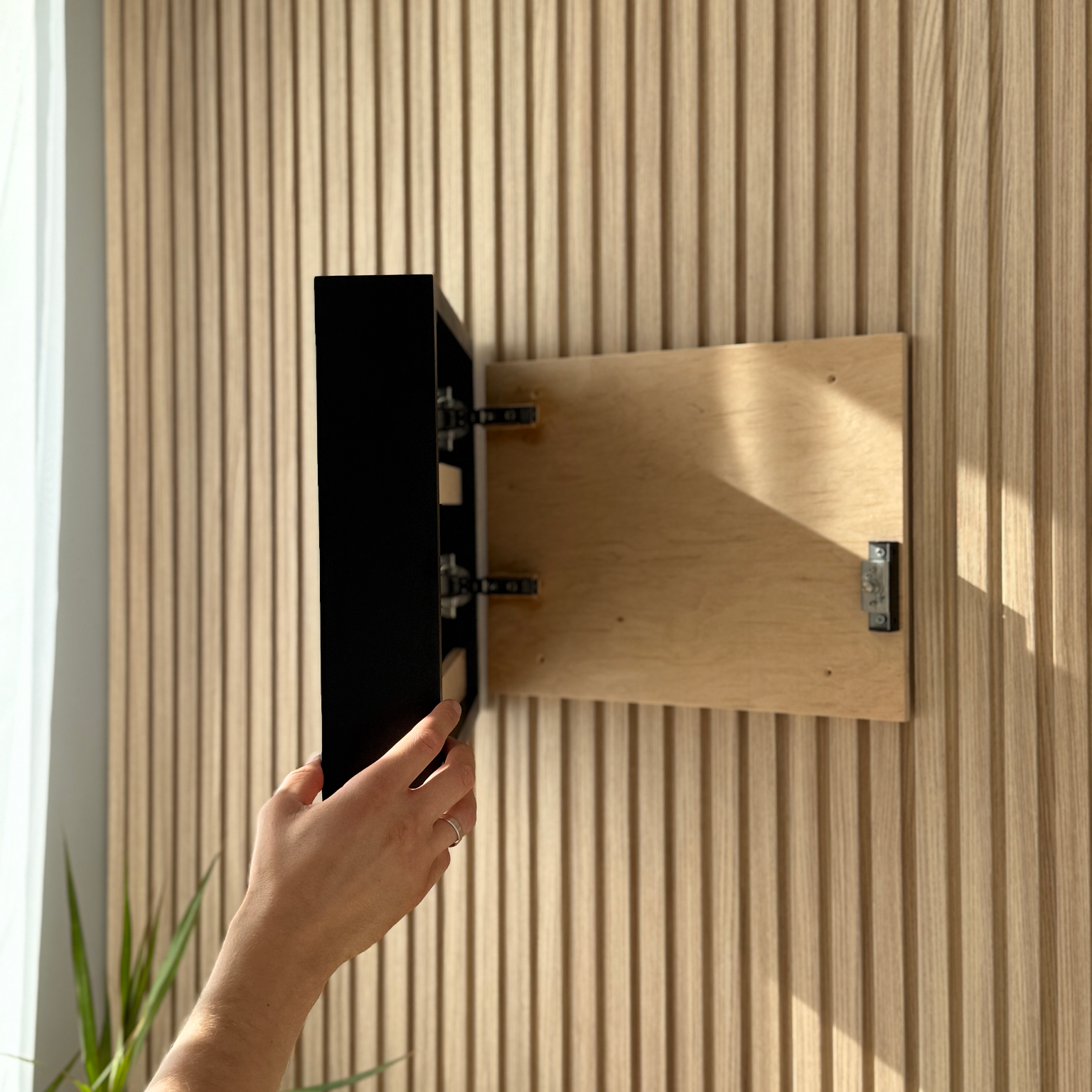 A hand is mounting a black wall-mounted cabinet onto a light wood slatted wall. The cabinet door is swung open to reveal the interior, and the scene is lit by natural sunlight with soft shadows.