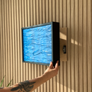 Hidden Wall Storage with Contactless RFID Lock. A person's tattooed arm is shown sliding a square black-framed textured blue artwork onto a light wood panel mounted on a wood slat wall. 