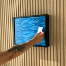 A framed wall-mounted artwork showing a blue textured water scene, mounted on a beige wood slat wall. A person's tattooed forearm and hand are visible holding a small white card in front of the frame. Hidden Wall Storage with Contactless RFID Lock