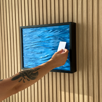 A framed wall-mounted artwork showing a blue textured water scene, mounted on a beige wood slat wall. A person's tattooed forearm and hand are visible holding a small white card in front of the frame. Hidden Wall Storage with Contactless RFID Lock