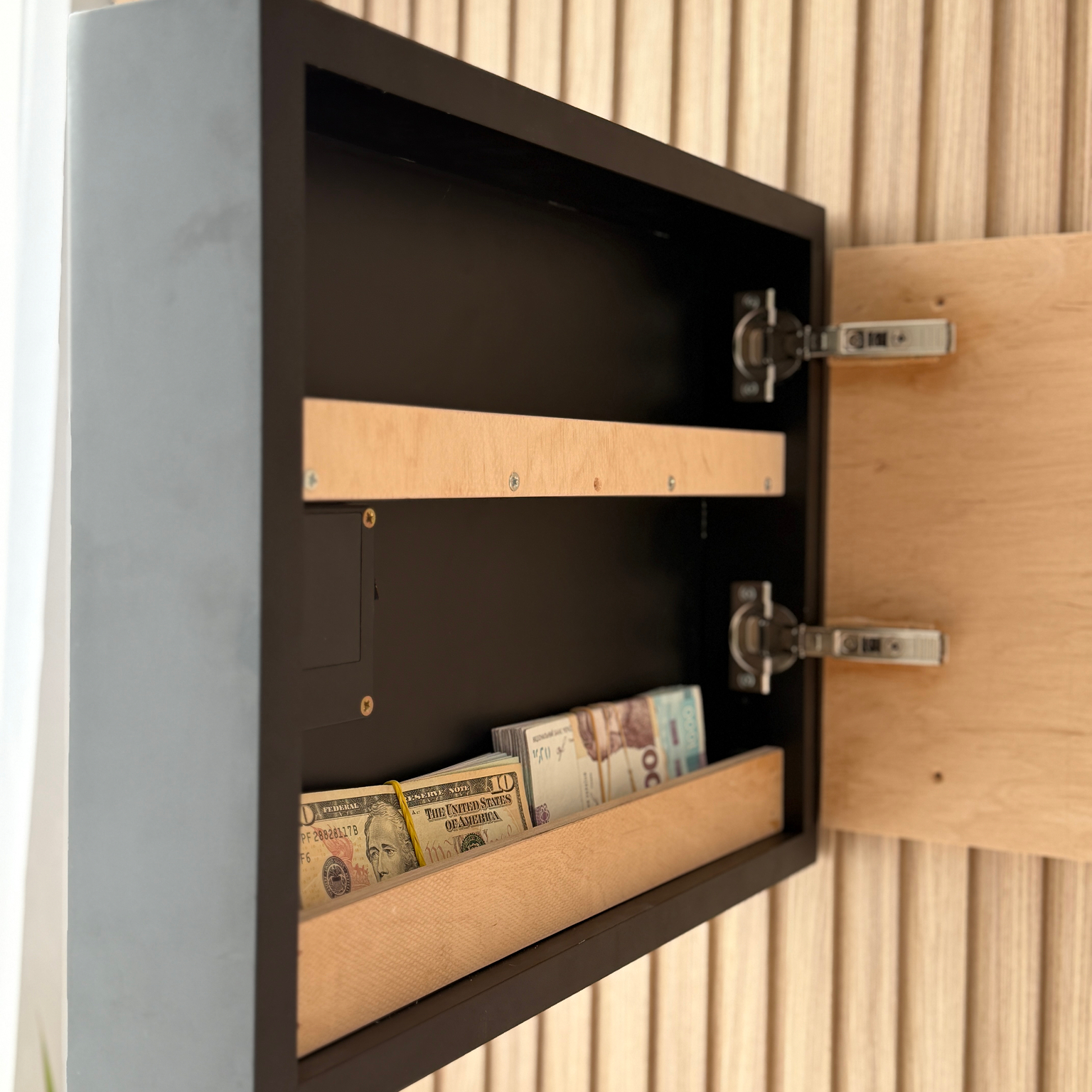 Wave Wall Art with Hidden RFID Storage Compartmentю A black wall-mounted safe or storage cabinet shown open, with wooden interior shelves and cash stored inside, set against a light wood slatted wall.
