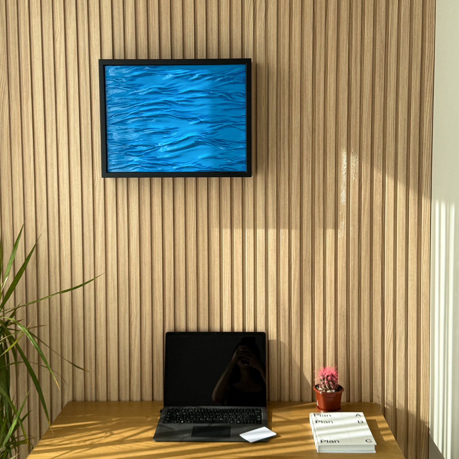 Wave Wall Art with Hidden RFID Storage Compartment. A framed abstract blue water-ripple artwork hangs on a light wood slatted wall above a wooden desk with an open laptop, a small potted pink plant, and a stack of books.