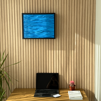 Wave Wall Art with Hidden RFID Storage Compartment. A framed abstract blue water-ripple artwork hangs on a light wood slatted wall above a wooden desk with an open laptop, a small potted pink plant, and a stack of books.