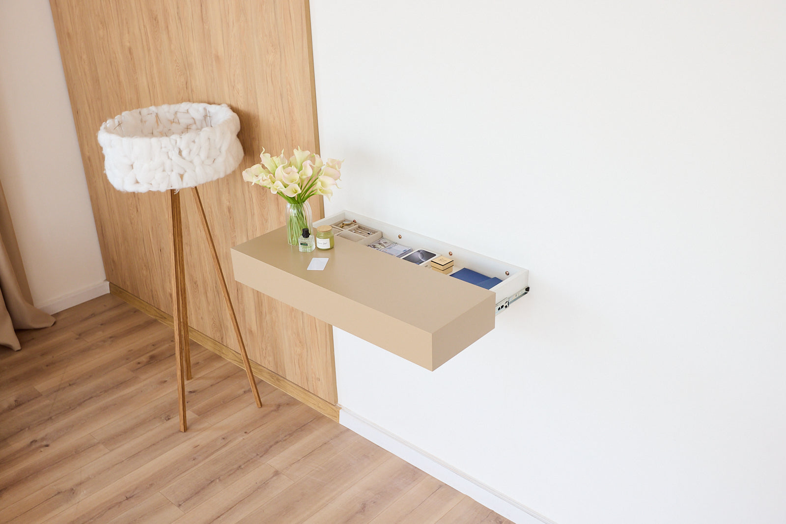 A minimalist wall-mounted desk in a light wood finish, shown partially pulled out against a white wall in a modern room with light wood flooring and a white floor lamp