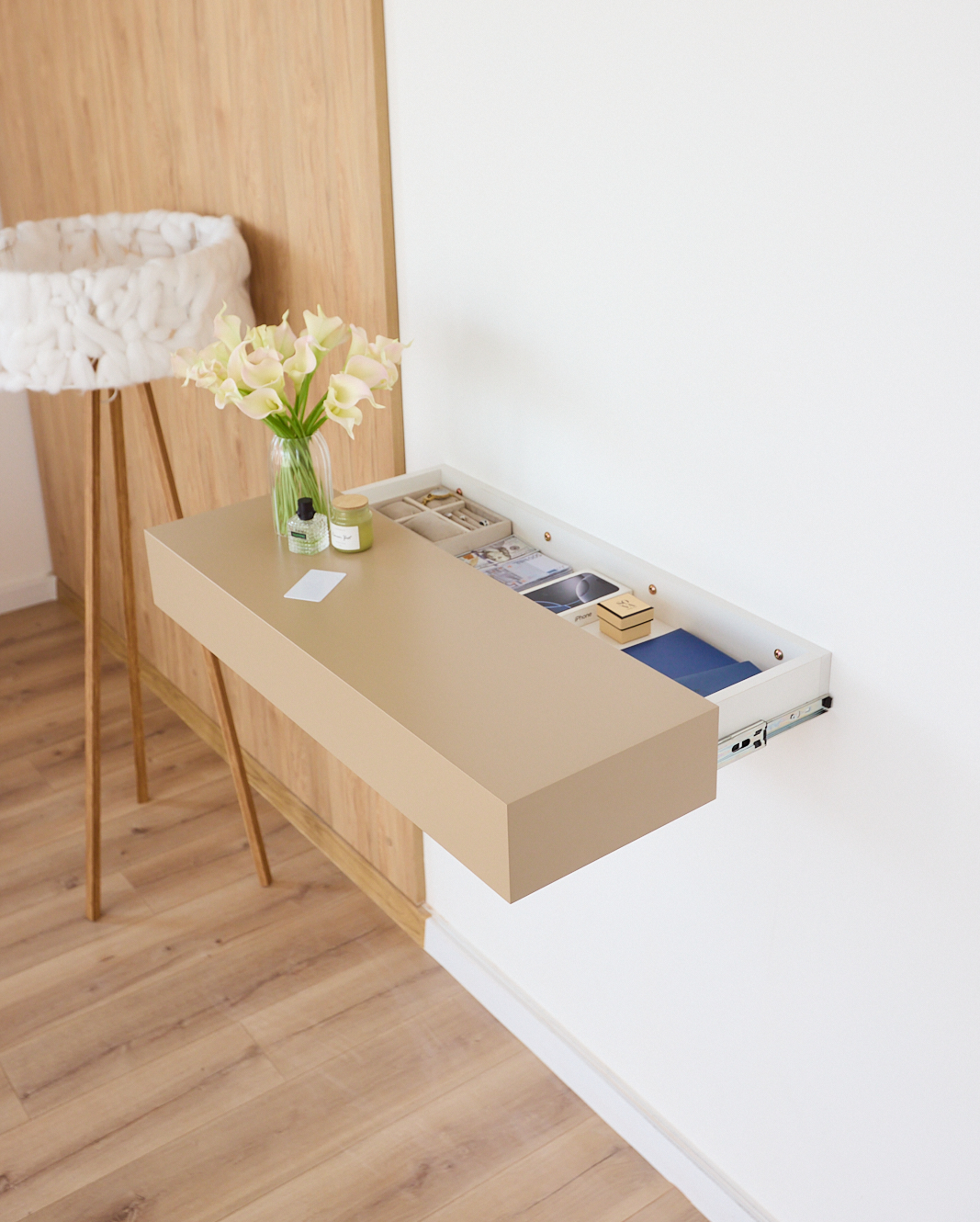 A wall-mounted floating desk in a warm beige wood finish with a white drawer compartment, shown installed on a white wall in a modern room with light wood flooring and a white floor lamp
