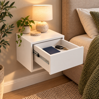 A modern white floating nightstand mounted on a beige wall beside a bed, with one drawer pulled open to show storage space. The nightstand is styled with a lit table lamp, a small potted plant, and a round coaster, set in a warm, cozy bedroom environment.