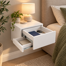A white floating wall-mounted nightstand drawer shown in a bedroom setting, with one drawer open to reveal storage space. The scene includes a lamp, small plant, pillows, and a textured blanket.