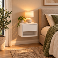 A modern white floating nightstand in a warm, styled bedroom setting next to a bed with neutral linens and a green knit throw, illuminated by a table lamp on top.